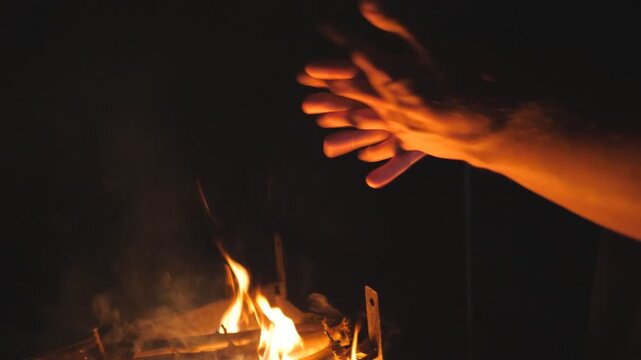 Guy warms his arms by a fire from metal brazier against the black background. Young man rubs his hands in front of an open bonfire from mangal at night. Concept of warmth and tranquil. Slow motion