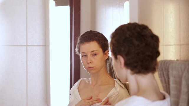 cancer patient looks at her reflection with a visible chest port after shower routine, expressing quiet concern, self monitoring and everyday oncology self care