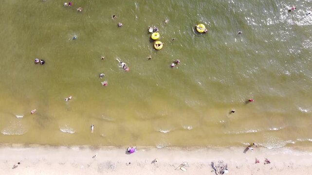 City beach park shoreline on Lake Granbury, swimmers clustered buoyed swim zone, sandy public waterfront along central recreation strip. Pale sand bands, sunlit bathers, open lakeside frame, TX