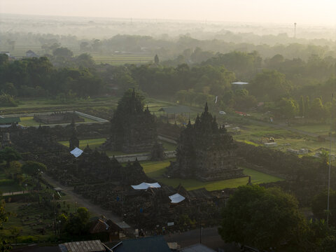 Aerial view of the ancient stone structures of Plaosan Lor Temple surrounded by morning mist and lush green trees in Kecamatan Prambanan, Jawa Tengah, Indonesia.