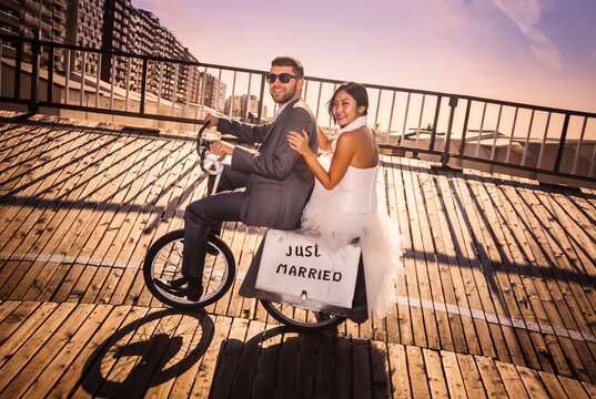 Playful wedding couple riding a vintage tandem bicycle on a seaside boardwalk, happy bride and groom having fun on a funny bike at a coastal beach resort.