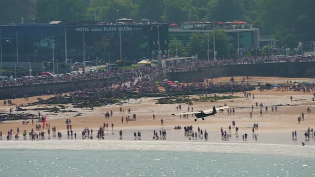 GIJON, SPAIN - JULY 27, 2025: Small propeller aircraft performing low pass over densely packed Gijon beach during Gijon International Air Festival