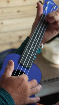 Ukulele music playing, man strumming blue instrument indoors, relaxed practice. Vertical video.