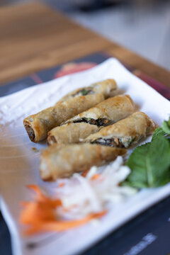 Fried spring rolls rest on a white plate. Thinly sliced carrots and radish add color. Fresh basil leaves garnish the dish. Dishes presented on a wooden table surface