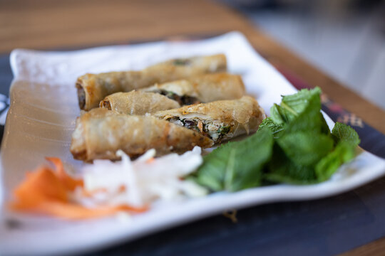 Fried spring rolls served on a white plate. Fresh mint and shredded vegetables add vibrant color. Delicate rolls showcase savory filling inside. Served with crisp greens and carrot garnish