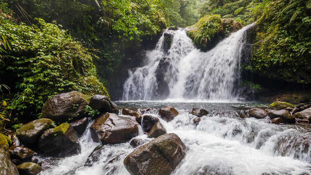 Aerial view of Ambyar waterfall cascading over dark mossy rocks surrounded by dense tropical forest in Purwokerto, Central Java, Indonesia.