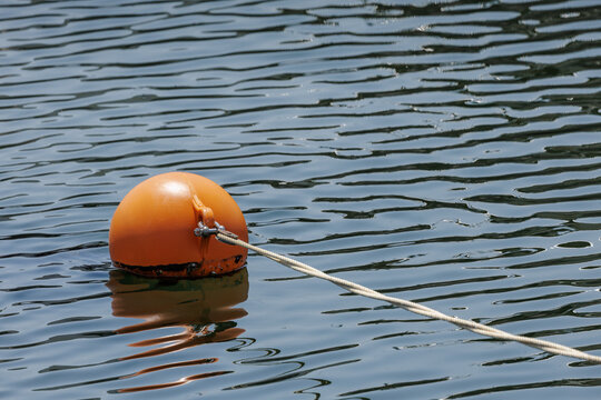 Orange mooring buoy tethered with white rope stretching into the distance in calm rippled water. Gentle surface waves shimmer in abstract patterns across the marina's basin.