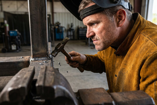 Side profile of mature craftsman with flipped-up helmet cleaning newly welded steel joint with pointed slag hammer, jaw clenched in deep professional concentration.