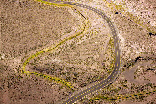 Aerial view of a winding asphalt road curving through a dry, rocky desert landscape with sparse green shrubs and arid terrain in Socaire, Antofagasta, Chile.