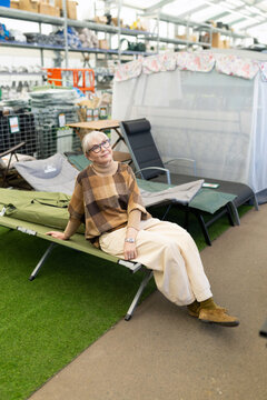 Mature woman sitting on a camping cot, exploring outdoor gear in a retail store