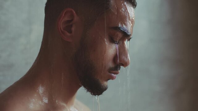 A man with dark skin stands with his eyes closed as water drips down his face. He is in a shower with a neutral background and a soft light. The focus is on his expression and the water.
