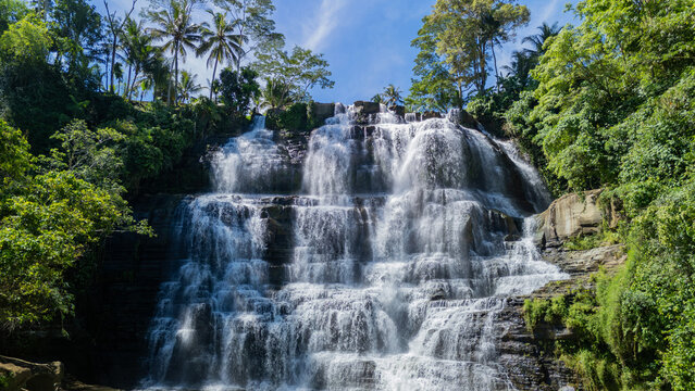 Aerial view of Cigangsa Waterfall cascading down tiered rock formations surrounded by lush tropical forest and palm trees in Sukabumi, West Java, Indonesia.