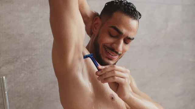A man with light skin is shaving his armpit in the shower. He appears happy and focused on his self-care routine. Water is running and steam is visible in the bathroom.