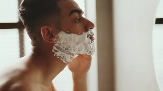 A man with medium skin tone is applying shaving cream on his face in front of a mirror. The setting is bright with natural light coming through a window.