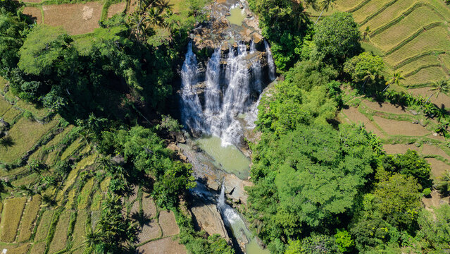 Aerial view of Cigangsa Waterfall cascading over dark rocks, surrounded by dense tropical forest and terraced rice paddies in Sukabumi, West Java, Indonesia.