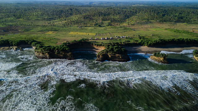 Aerial view of Ujung Genteng Beach featuring rugged rocky cliffs, crashing ocean waves, and lush green coastal vegetation in Sukabumi, West Java, Indonesia.