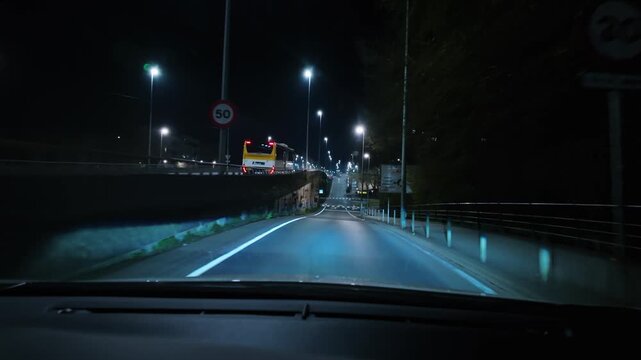 Point of view shot from a car driving on a quiet urban street, following a bus through roundabouts and underpasses illuminated by bright streetlights during the night in the city