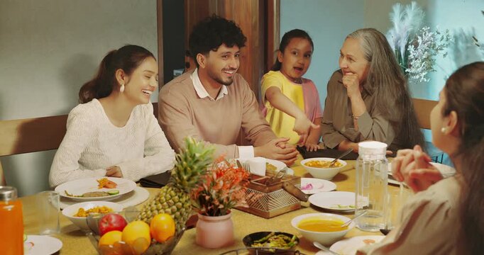 Indian happy relax joint family group sitting around table eat food talk speak share gossip have fun joy Young adult male female aged mom girl boy child spend time enjoy night dinner room home place