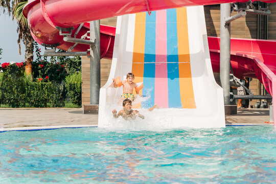 Two Childs boys laughing while slide in pool after going down water slide at sunny day in water Aqua park. Kids refreshing at heat weather, active vacation and healthy lifestyle. Happy summer.