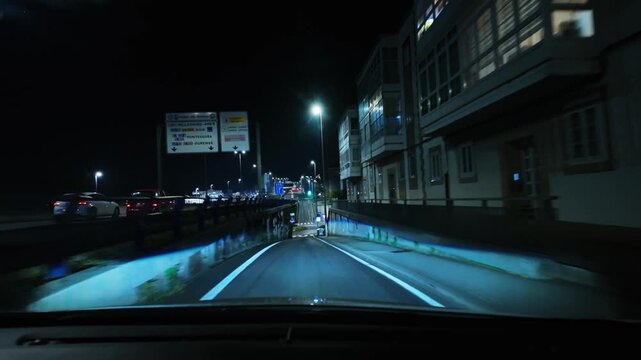 Point of view shot from inside a car driving through an illuminated city at night, passing traffic lights, street signs, intersections, roundabouts, and other vehicles on the road