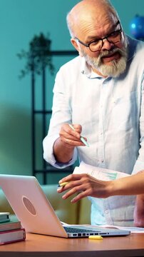 Indian young son and senior father filing online tax return together using laptop while standing at table in modern home, discussing financial reports, income statements yearly accounting documents