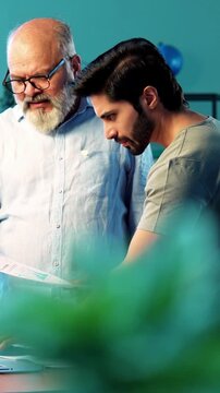 Indian young son and senior father filing online tax return together using laptop while standing at table in modern home, discussing financial reports, income statements yearly accounting documents