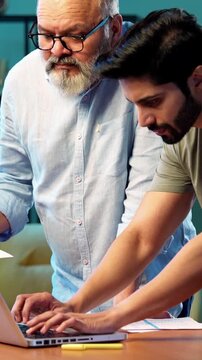 Indian young son and senior father filing online tax return together using laptop while standing at table in modern home, discussing financial reports, income statements yearly accounting documents