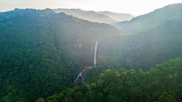 Aerial view of Cikanteh Waterfall cascading down a rocky cliff into a lush tropical valley surrounded by misty forested hills in Sukabumi, West Java, Indonesia.
