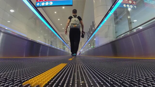 Ground-level tracking shot of a person with backpack walking on ribbed airport travelator, blue LED strip lights, glass walls, and modern architecture. (159)
