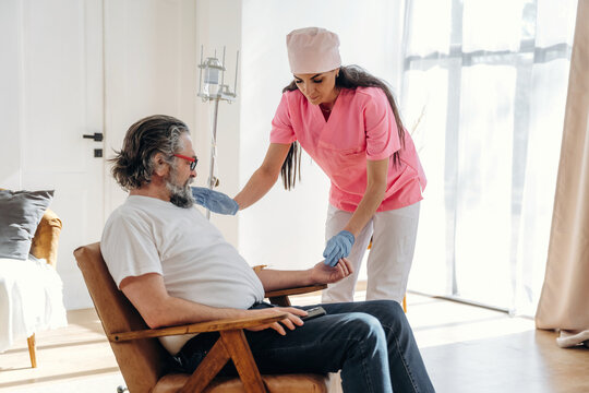 Female healthcare worker in pink scrubs administers care to elderly male patient with gray hair and glasses seated in a bright, modern indoor environment