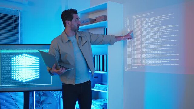 A professional male software engineer stands presenting and explaining lines of code projected on a wall display during a technical meeting, gesturing towards the data script in a dark room with drama