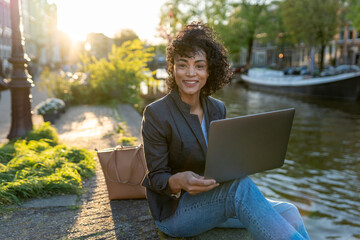 Woman using laptop for remote work by an amsterdam canal, symbolizing freelance, digital nomad, and travel freedom concepts © CatalnIlie