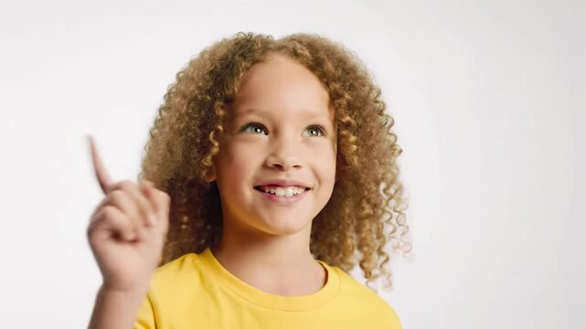 A cheerful young girl, curly hair & yellow t-shirt, has an intelligent eureka moment, raising her finger in a bright white studio, soft lighting. Concept of discovery