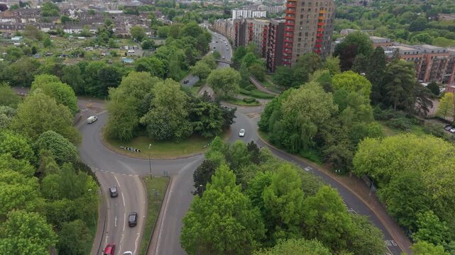Roundabout with traffic and modern apartment buildings in Maidstone, Kent, UK. Aerial view flying above the road with cars in motion. Pedestrian bridge crossing road towards green area with allotments