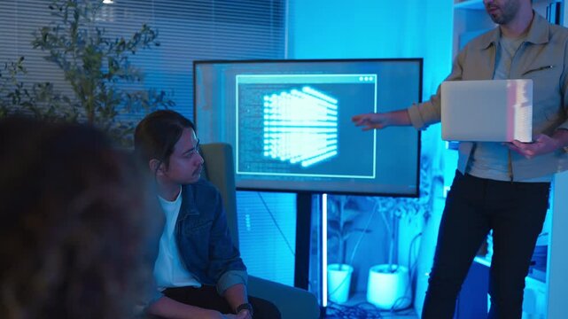A professional East Asian female software engineer stands holding a laptop and gesturing toward lines of code projected on a wall display, presenting data scripts to her multiethnic colleagues during 