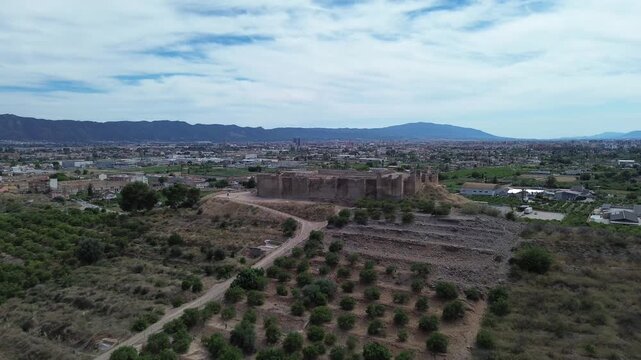 Aerial view of the historic islamic Castillejo de Monteagudo ruins surrounded by lush agricultural fields and majestic mountains under a bright cloudy blue sky during a sunny summer day