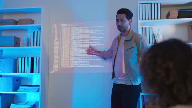 A professional male software engineer stands presenting and explaining lines of code projected on a wall display during a technical meeting, gesturing towards the data script in a dark room with drama