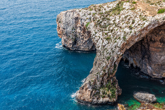 Landscape shot of side of the blue grotto malta natural cave archway with deep blue Mediterranean Sea water in summer with chalky limestone cliffs and lush green shrubs at sunset