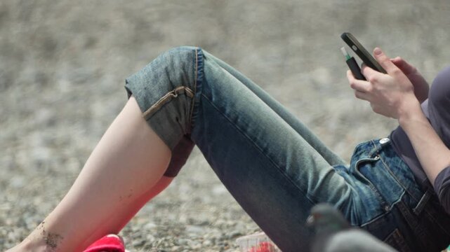 Woman smartphone beach young female sitting on pebble shore using mobile phone and holding electronic cigarette during summer vacation