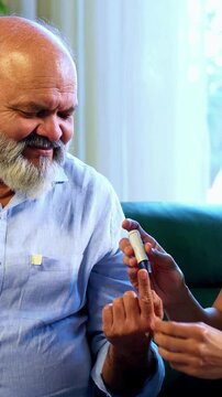 Indian caring young son checking blood sugar level of aged father using digital glucometer while sitting on sofa in modern home, concerned about glucose reading and supporting elderly parent