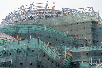 Fototapeta premium Construction of a large building in an urban area during cloudy weather with scaffolding and green netting present