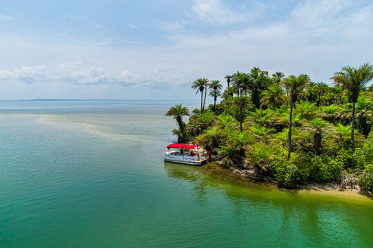 Aerial view of a boat with a red canopy docked at a lush tropical shoreline with palm trees and turquoise water in Monrovia, Liberia.