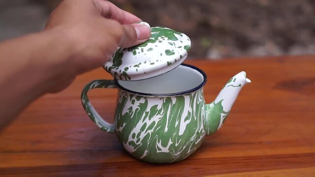 A hand is opening the lid of a green enamel Blirik teapot.