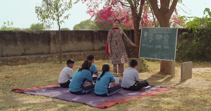 Indian young adult female tutor hold stick pointing words teach to girl boy under tree rural area place village group sitting on mat look board talk speak read do study learn open ground class field