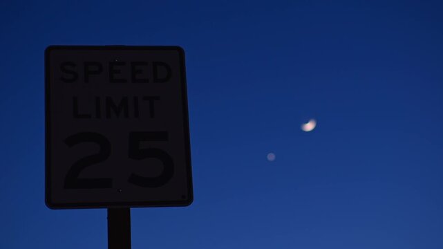 Speed limit sign against crescent moon in evening sky standing against a deep blue backdrop creating an unusual combination of urban infrastructure and celestial scenery