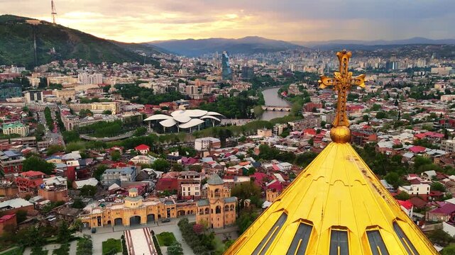 Aerial drone shot of the golden dome and upper architecture of the Holy Trinity Cathedral (Sameba) in Tbilisi, Georgia, with a panoramic view of the city and surrounding mountains in the background