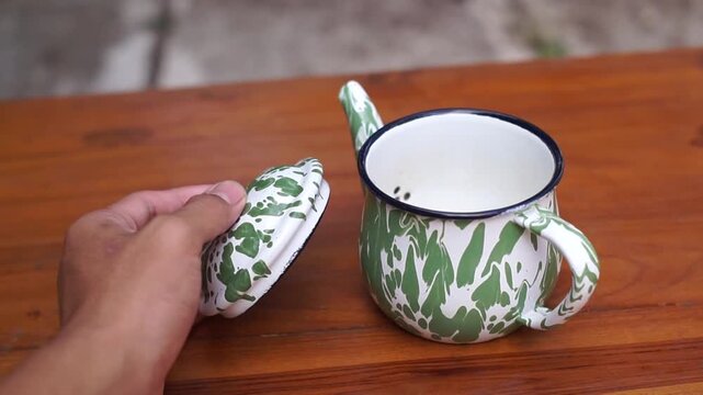 A hand is opening the lid of a green enamel Blirik teapot.