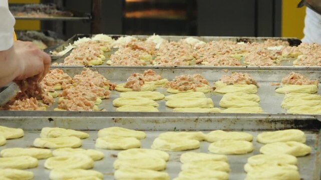 Bakery workers place savory filling on rows of raw pastry dough arranged on metal baking trays in a professional kitchen.
