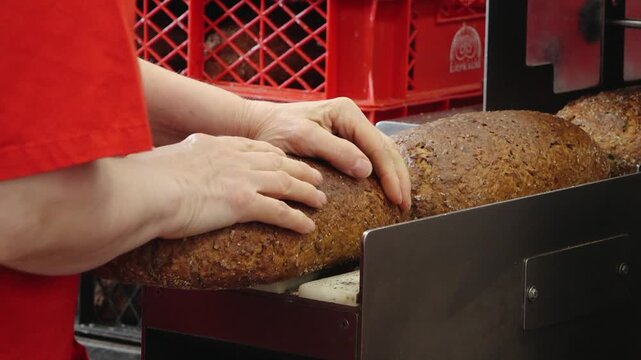 A bare hand steadies a large crusty loaf of dark bread on bakery slicing equipment, with red crates blurred in the background.