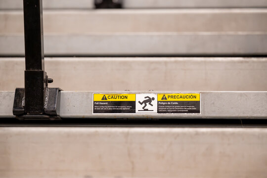 A bilingual English and Spanish caution label warning of fall hazard is affixed to the metal edge of bleacher steps, its yellow warning triangles and falling figure icon clearly legible.